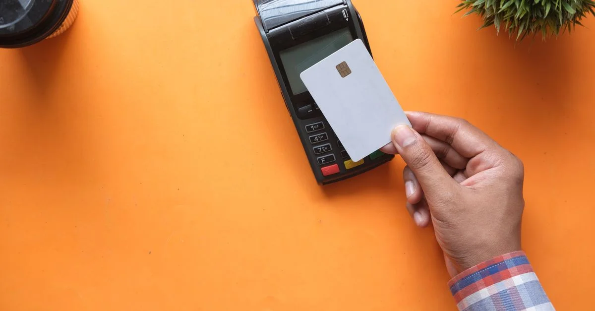 Une personne effectuant un paiement sans contact avec une carte de crédit et un lecteur de carte sur une surface orange vif.