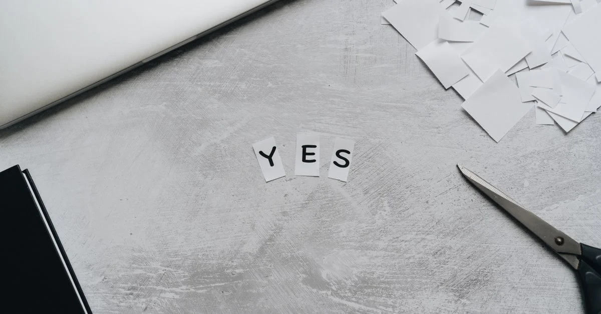 Composition artistique de découpes en papier formant 'OUI' avec des ciseaux sur une surface de bureau.