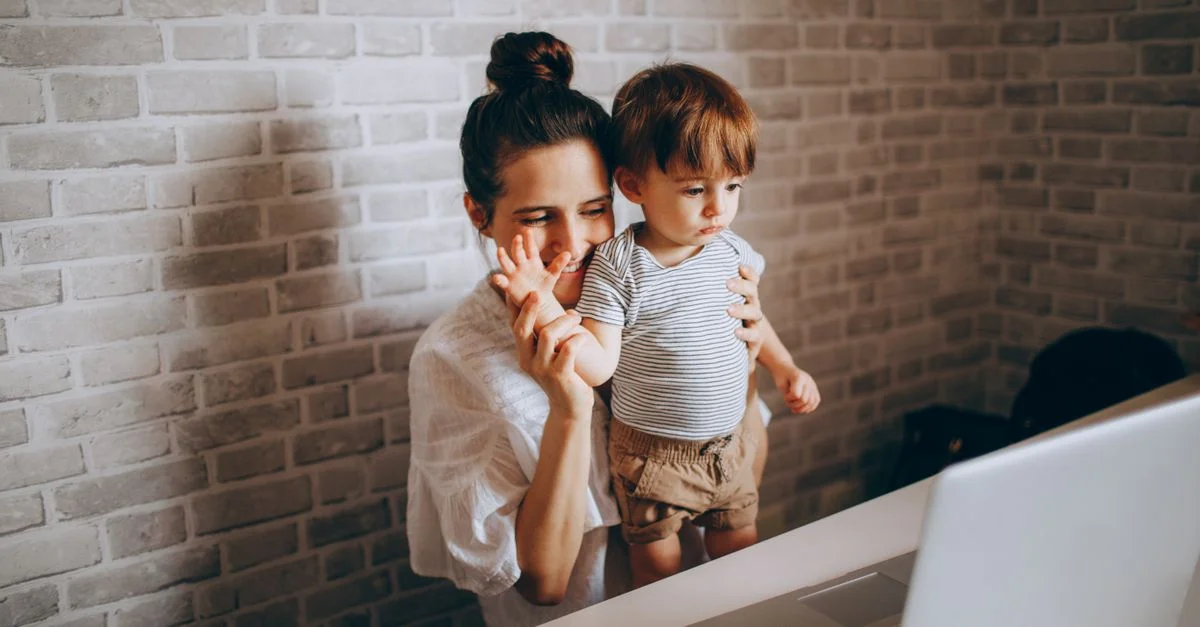 Une mère fait signe à un ordinateur portable avec son jeune enfant, souriant chaleureusement dans le cadre confortable de leur maison.
