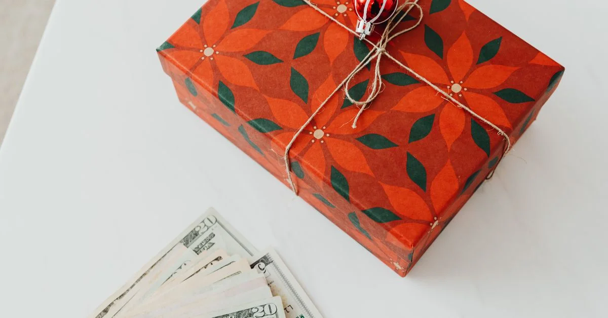 Boîte cadeau sur le thème de Noël, attachée avec une corde et des billets de banque à proximité sur une surface blanche.