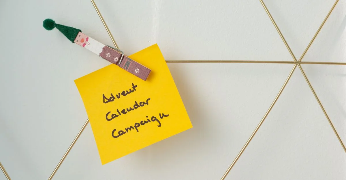 Note jaune avec 'Campagne du Calendrier de l'Avent' accrochée à une grille en fil géométrique, évoquant la planification des fêtes.