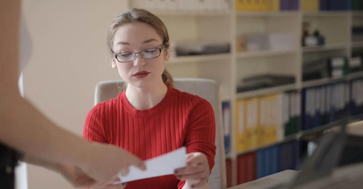 Femme concentrée en pull rouge révisant un document dans un environnement de bureau occupé.