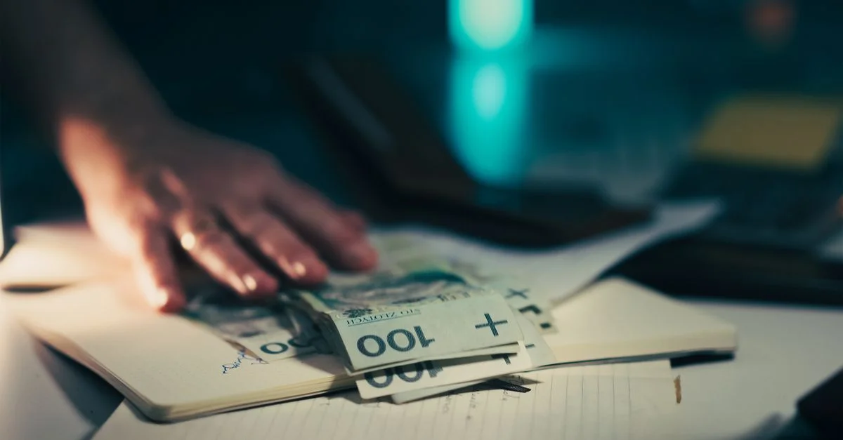 Gros plan sur une main qui se tend vers des billets de banque posés sur un bureau où se trouvent un ordinateur portable et une calculatrice.