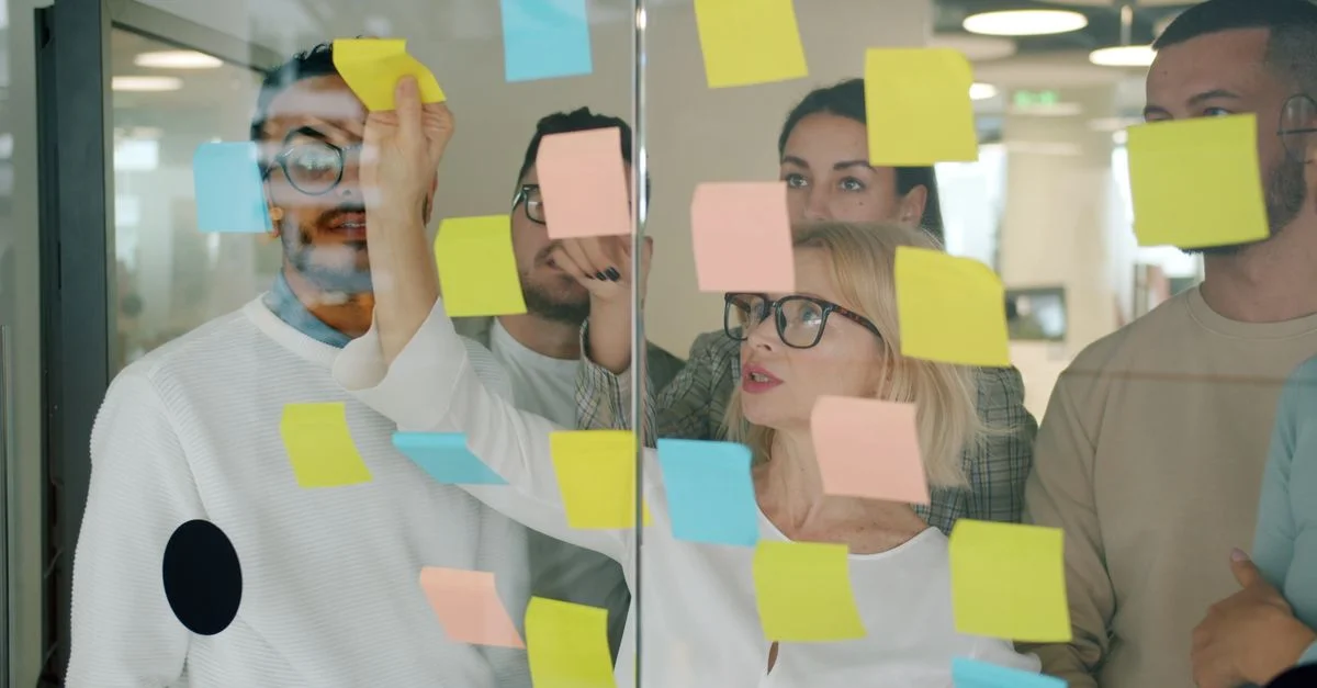 Une équipe aux profils variés se réunit pour un brainstorming à l'aide de post-it colorés sur une vitre dans un bureau moderne.