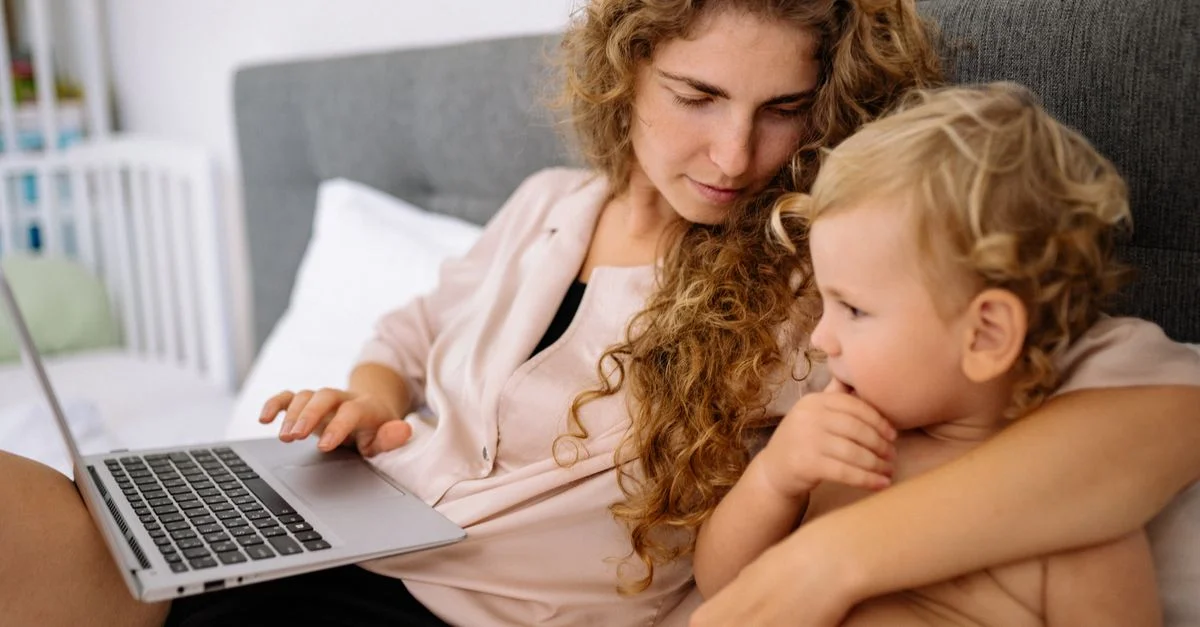 Une mère avec les cheveux blonds bouclés est assise avec son fils tout-petit sur un lit, utilisant un ordinateur portable.