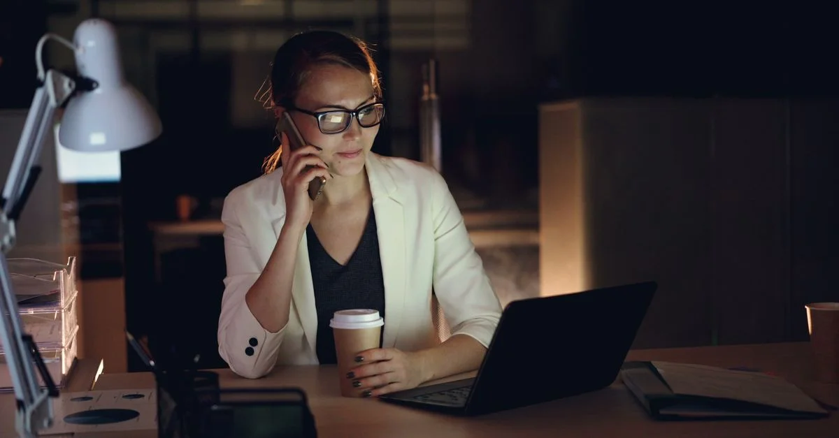 Femme en lunettes travaillant tard dans la nuit sur un ordinateur portable, tenant un café, en train de passer un appel téléphonique.