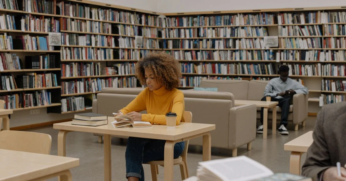 Étudiants étudiant dans une bibliothèque universitaire entourée d'étagères et de tables.